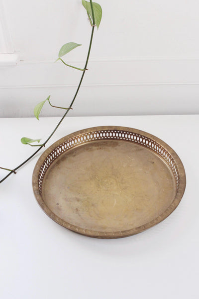 May include: A round, antique brass tray with a decorative pierced edge. The tray has a slightly worn appearance, suggesting age and use. A green plant stem with leaves is in the background, adding a touch of nature.