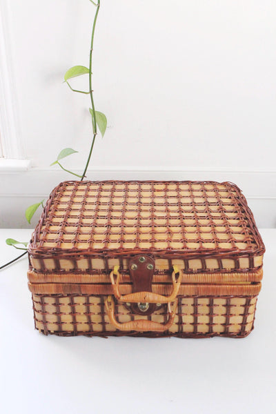 May include: A rectangular, vintage picnic basket made of woven brown and tan wicker. It features a leather handle and clasp, with a woven pattern on the lid and sides. The basket is designed for carrying food and drinks.