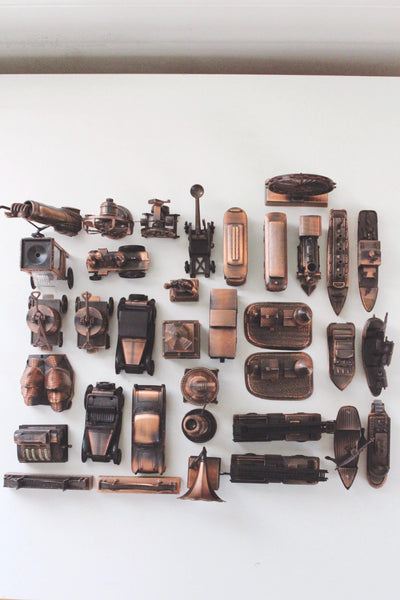 May include: A collection of vintage-style metal pencil sharpeners in various shapes, including cars, boats, and cannons. The sharpeners are in shades of bronze and copper, arranged against a white background.