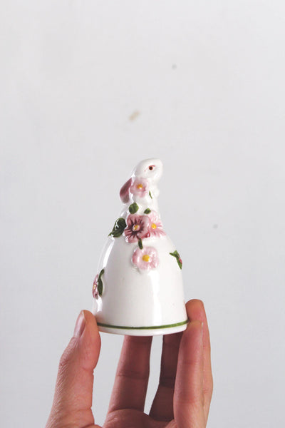 May include: A white ceramic bell-shaped figurine with a bunny head and pink flower accents. The figurine has a green base and is held against a white background. The bunny has pink ears and red eyes.