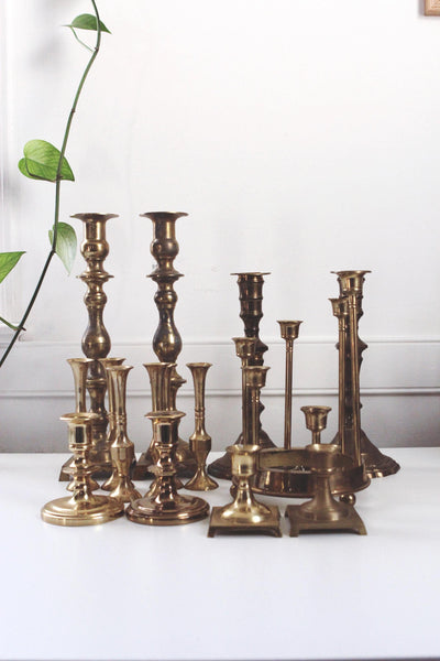 May include: A collection of antique brass candlesticks in various shapes and sizes. The candlesticks have a warm, golden hue and are arranged on a white surface. Some have ornate details, while others are more minimalist.